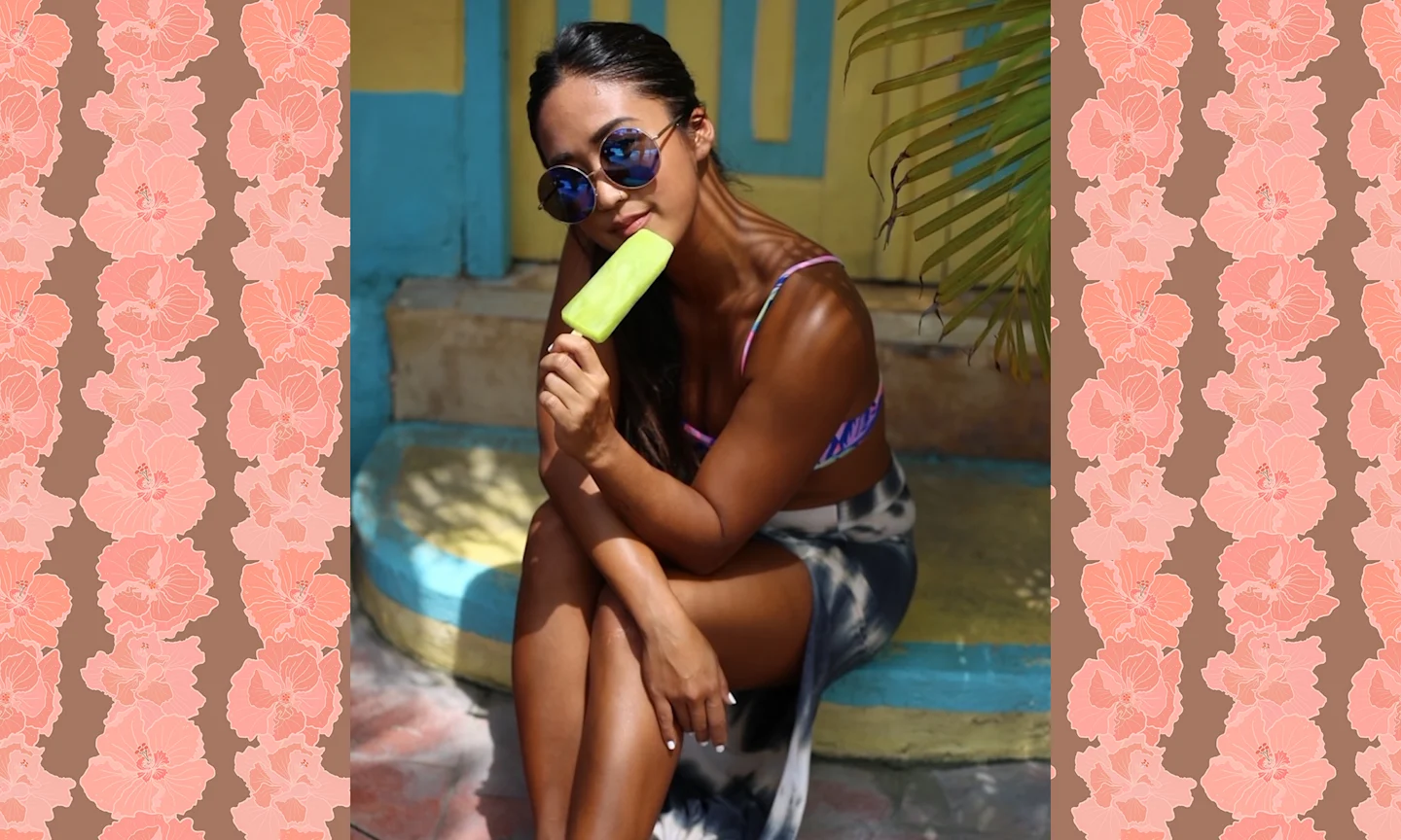 a woman in a swimsuit is sitting with her legs crossed. She is wearing sunglasses and holding a green popsicle.